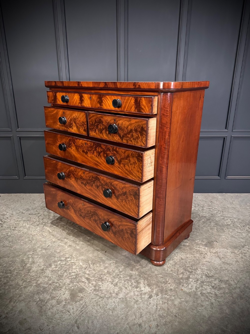 A tall Victorian flame mahogany chest of drawers featuring a decorative top drawer, five lower drawers with dark turned handles, rounded pilasters, and a solid plinth base. View IX