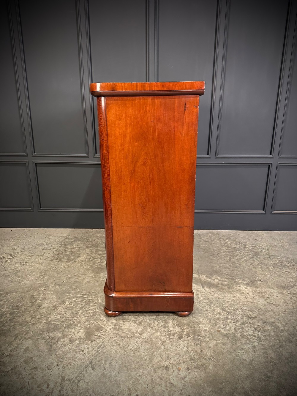 A tall Victorian flame mahogany chest of drawers featuring a decorative top drawer, five lower drawers with dark turned handles, rounded pilasters, and a solid plinth base. View X