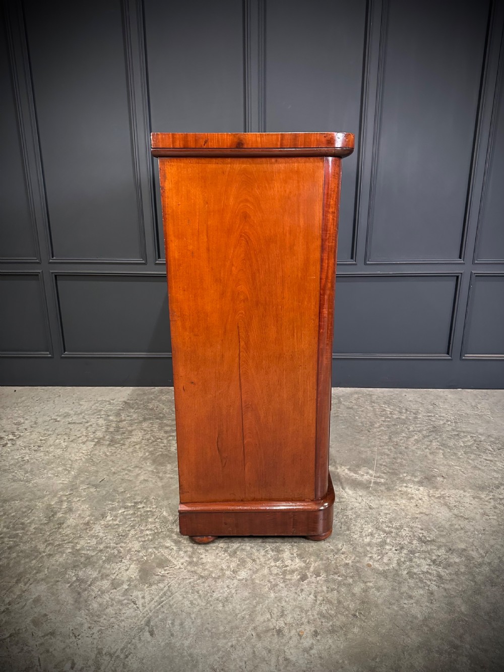 A tall Victorian flame mahogany chest of drawers featuring a decorative top drawer, five lower drawers with dark turned handles, rounded pilasters, and a solid plinth base. View XII