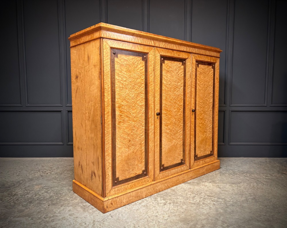 A low Victorian three-door wardrobe crafted from birds eye maple featuring dark wood inlaid borders and turned wooden knobs.