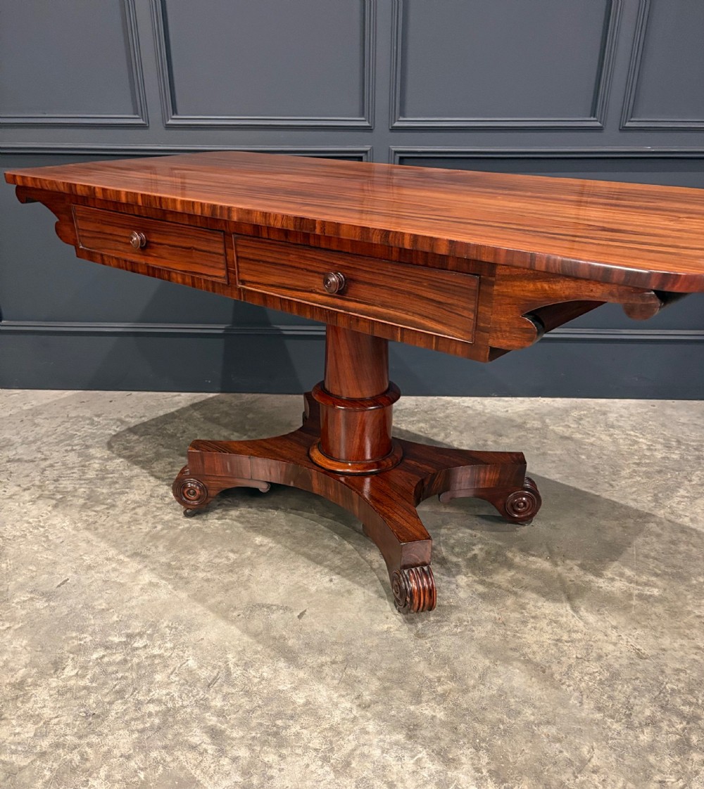 A 19th-century Goncalo Alves library table featuring a polished rectangular top with rounded corners, frieze drawers, and a central turned pedestal on a quadform base with scroll feet. View VIII