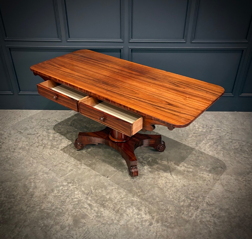 A 19th-century Goncalo Alves library table featuring a polished rectangular top with rounded corners, frieze drawers, and a central turned pedestal on a quadform base with scroll feet. View IX