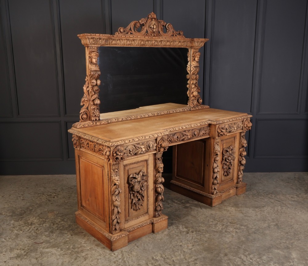 A large bleached oak pedestal sideboard featuring elaborate fruit swags, carved mask details, twin lower cupboards, and an integrated rectangular mirror superstructure.