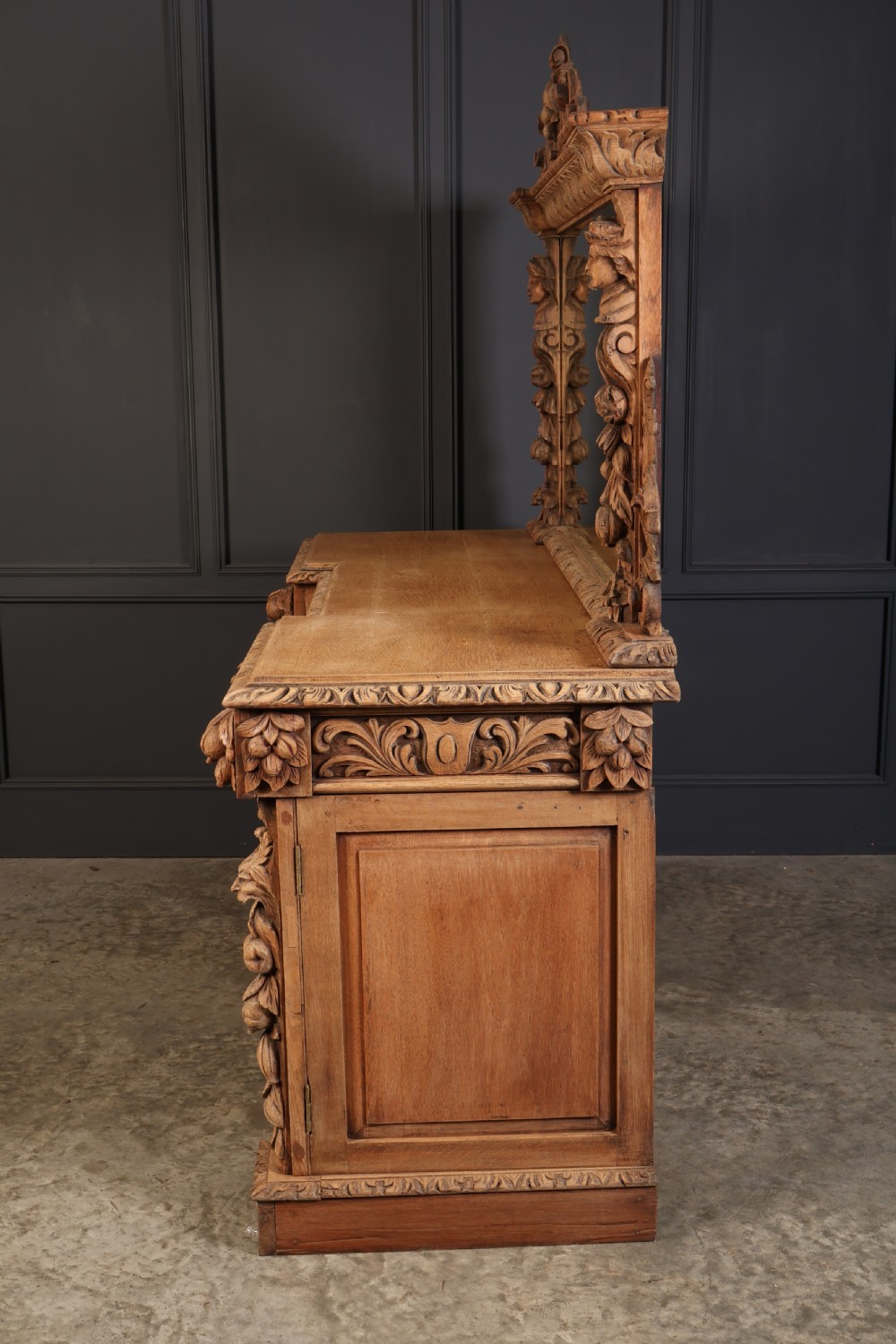 A large bleached oak pedestal sideboard featuring elaborate fruit swags, carved mask details, twin lower cupboards, and an integrated rectangular mirror superstructure. View VII