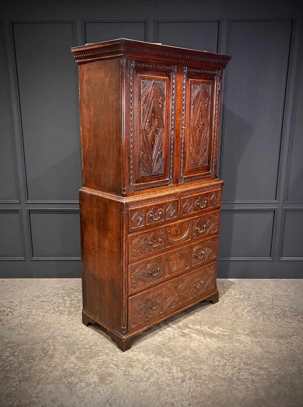 An 18th-century carved dark oak housekeepers cupboard featuring upper panel doors with geometric lozenge carving above a base with five carved drawers. View II