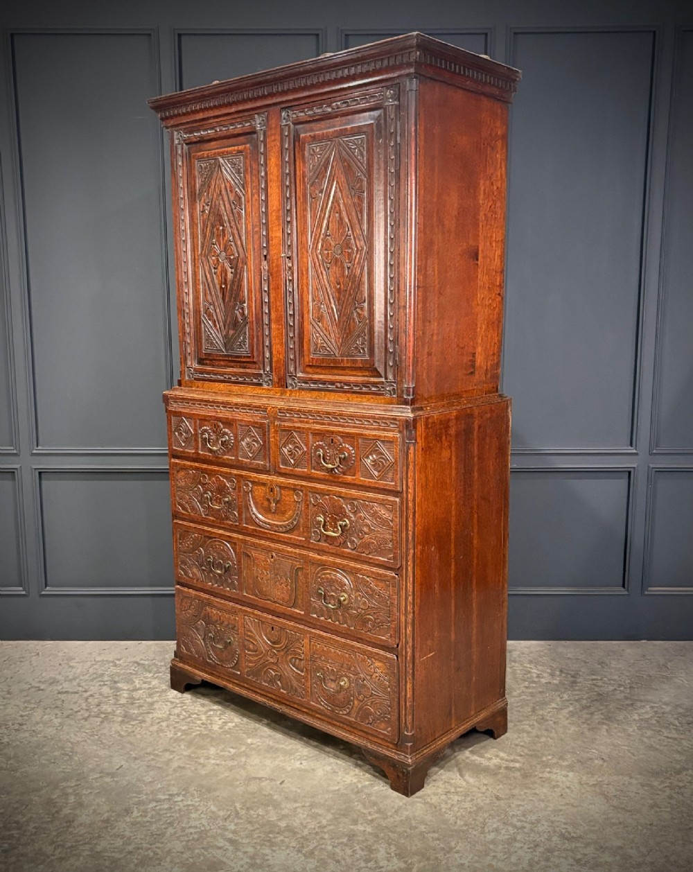 An 18th-century carved dark oak housekeepers cupboard featuring upper panel doors with geometric lozenge carving above a base with five carved drawers.
