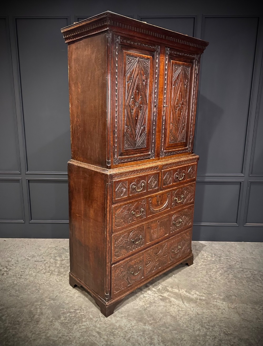 An 18th-century carved dark oak housekeepers cupboard featuring upper panel doors with geometric lozenge carving above a base with five carved drawers. View XII