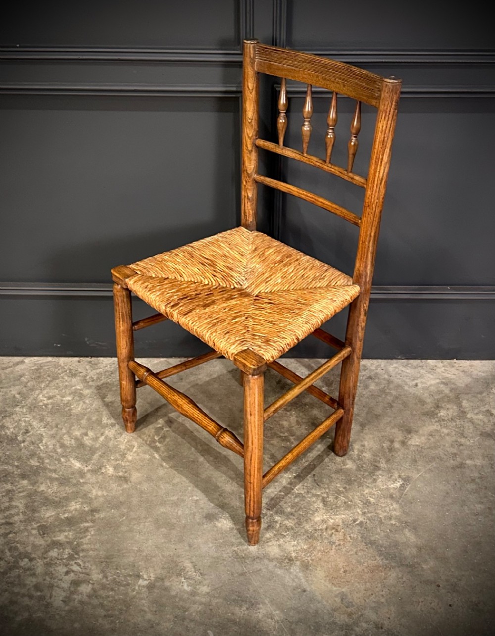An 18th-century country elm side chair featuring a woven rush seat, turned spindle backrest, and turned wood legs connected by stretchers. View V