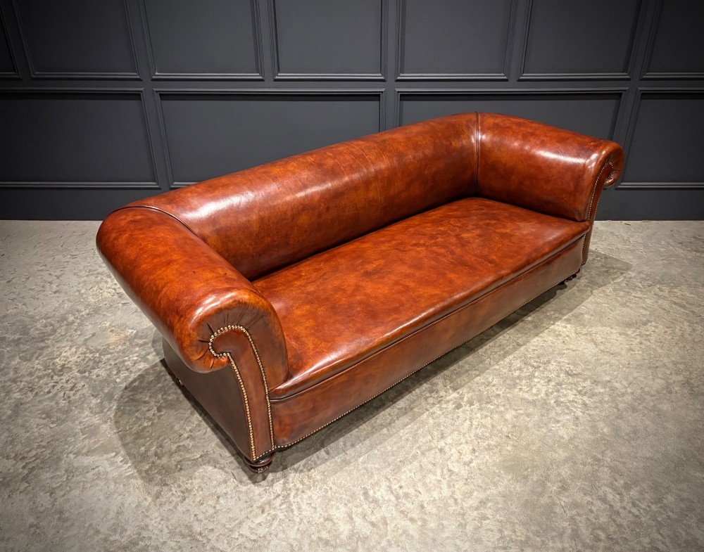 A Victorian chestnut brown leather sofa featuring continuous rolled arms, brass studding detail, and turned wood legs on castors. View III