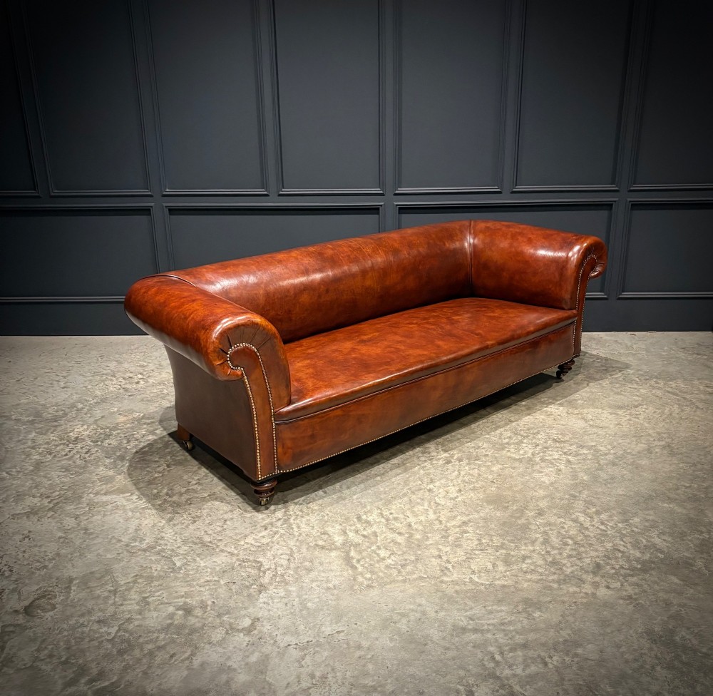 A Victorian chestnut brown leather sofa featuring continuous rolled arms, brass studding detail, and turned wood legs on castors. View IV
