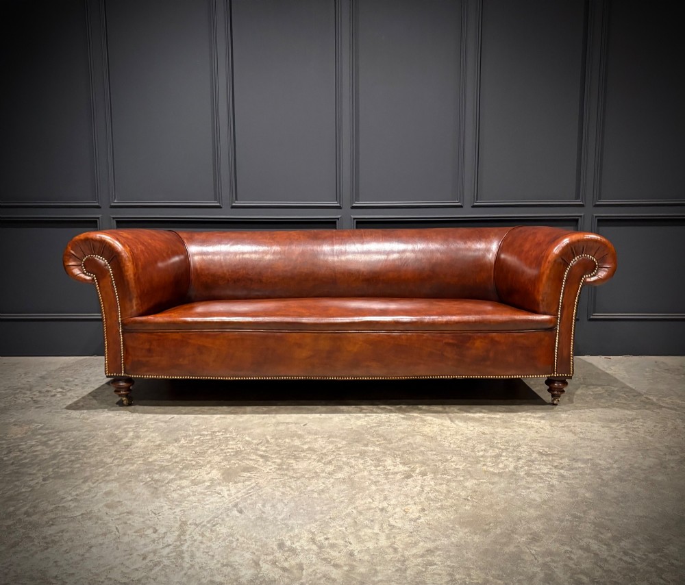 A Victorian chestnut brown leather sofa featuring continuous rolled arms, brass studding detail, and turned wood legs on castors.