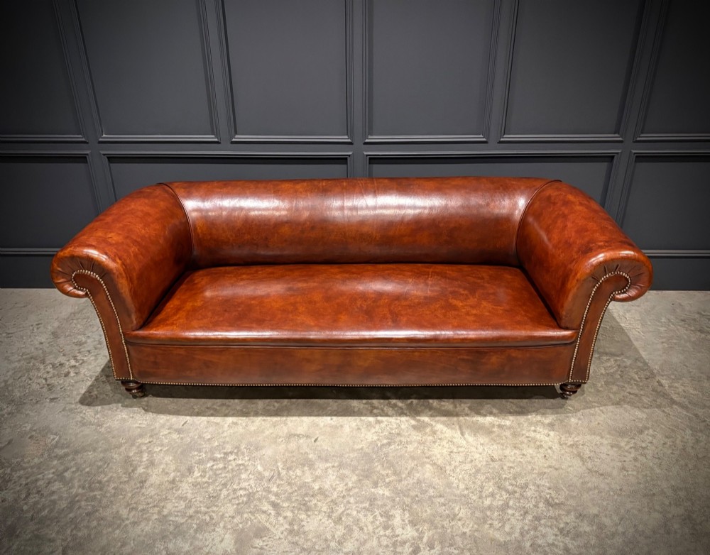 A Victorian chestnut brown leather sofa featuring continuous rolled arms, brass studding detail, and turned wood legs on castors. View V