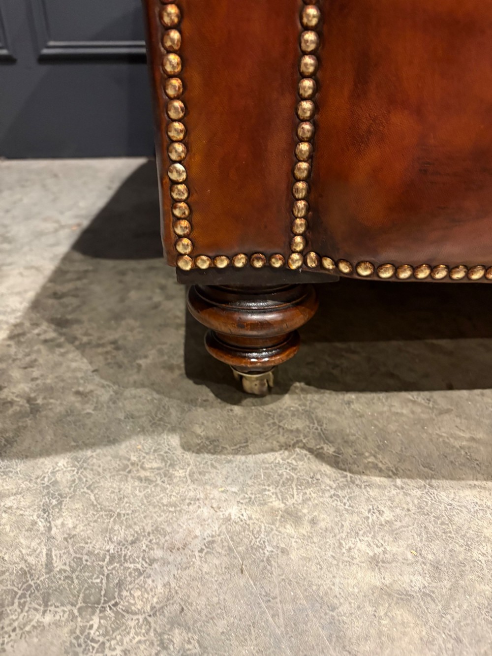 A Victorian chestnut brown leather sofa featuring continuous rolled arms, brass studding detail, and turned wood legs on castors. View VI