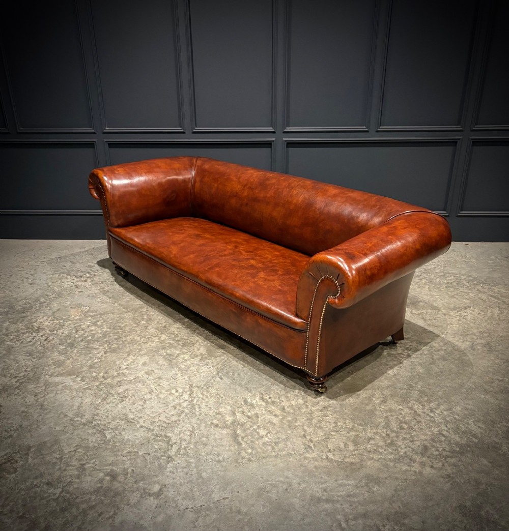 A Victorian chestnut brown leather sofa featuring continuous rolled arms, brass studding detail, and turned wood legs on castors. View VII