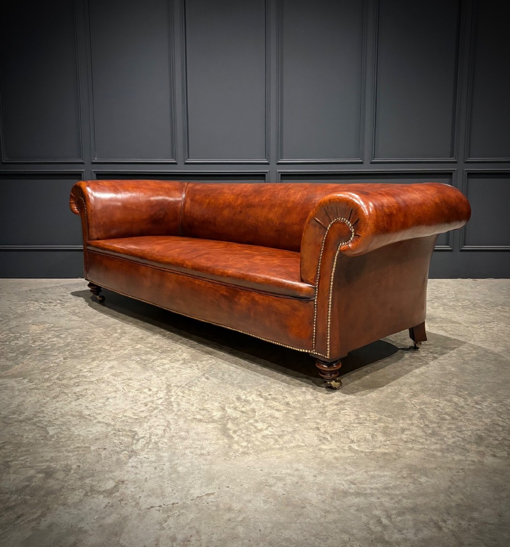 A Victorian chestnut brown leather sofa featuring continuous rolled arms, brass studding detail, and turned wood legs on castors. View VIII