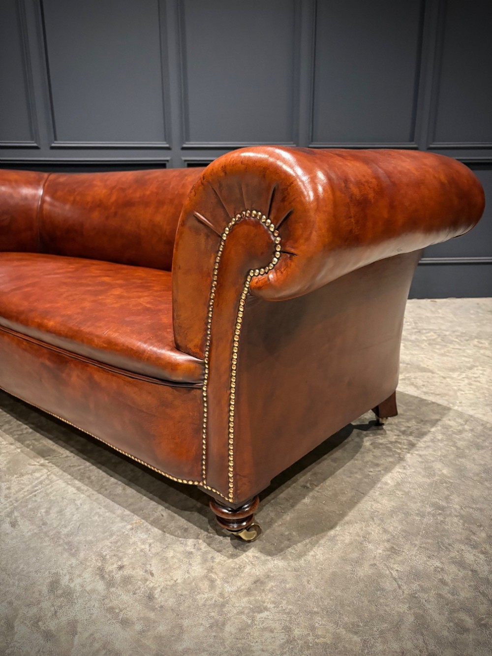 A Victorian chestnut brown leather sofa featuring continuous rolled arms, brass studding detail, and turned wood legs on castors. View IX