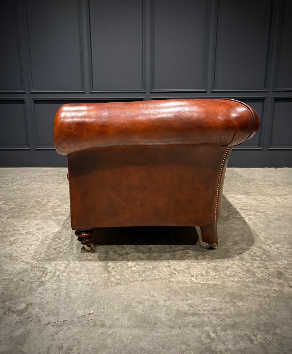 A Victorian chestnut brown leather sofa featuring continuous rolled arms, brass studding detail, and turned wood legs on castors. View X