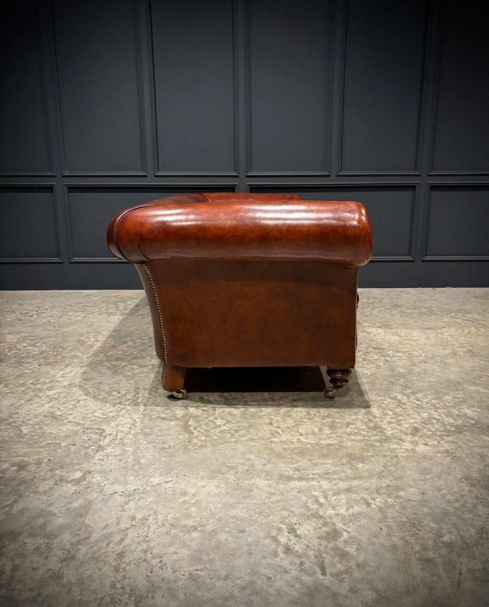 A Victorian chestnut brown leather sofa featuring continuous rolled arms, brass studding detail, and turned wood legs on castors. View XII