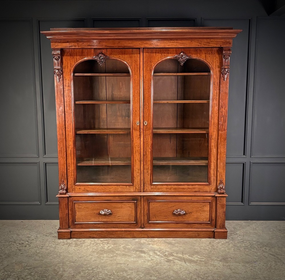 A large Victorian mahogany bookcase featuring two arched glazed doors, carved corbels, internal wooden shelves, and two drawers at the base. View II