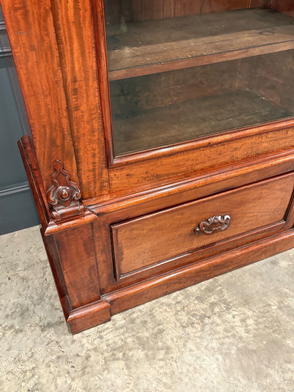 A large Victorian mahogany bookcase featuring two arched glazed doors, carved corbels, internal wooden shelves, and two drawers at the base. View IV