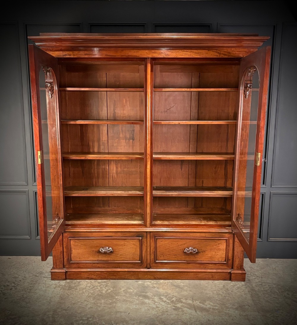 A large Victorian mahogany bookcase featuring two arched glazed doors, carved corbels, internal wooden shelves, and two drawers at the base. View V