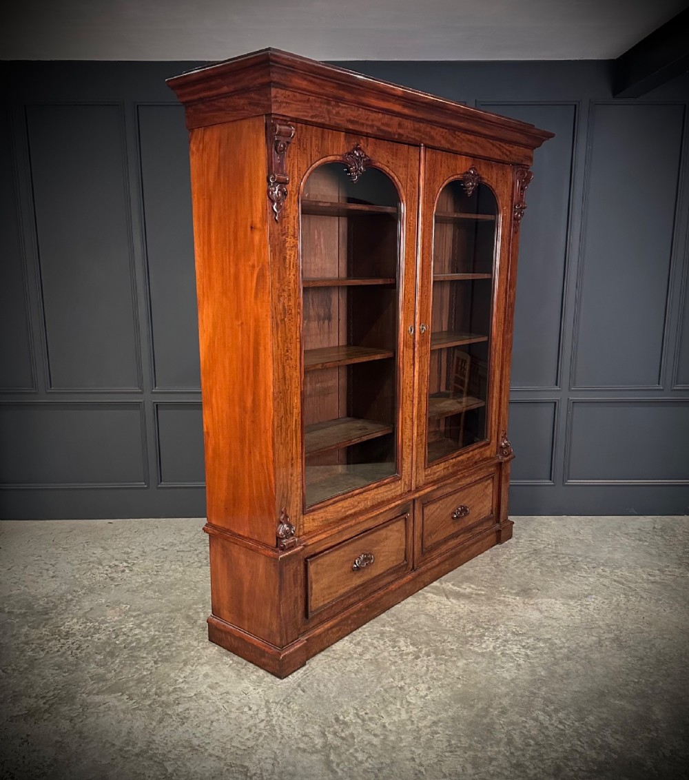 A large Victorian mahogany bookcase featuring two arched glazed doors, carved corbels, internal wooden shelves, and two drawers at the base. View IX