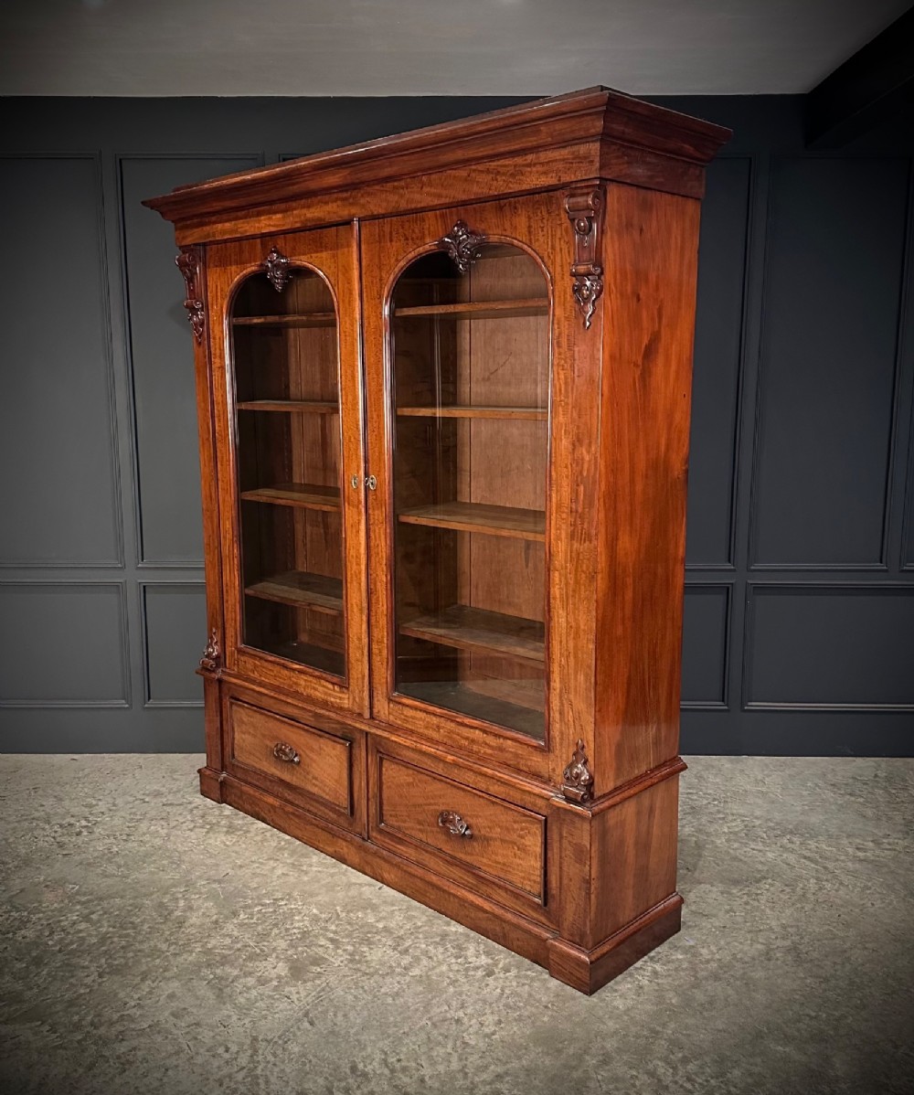 A large Victorian mahogany bookcase featuring two arched glazed doors, carved corbels, internal wooden shelves, and two drawers at the base.