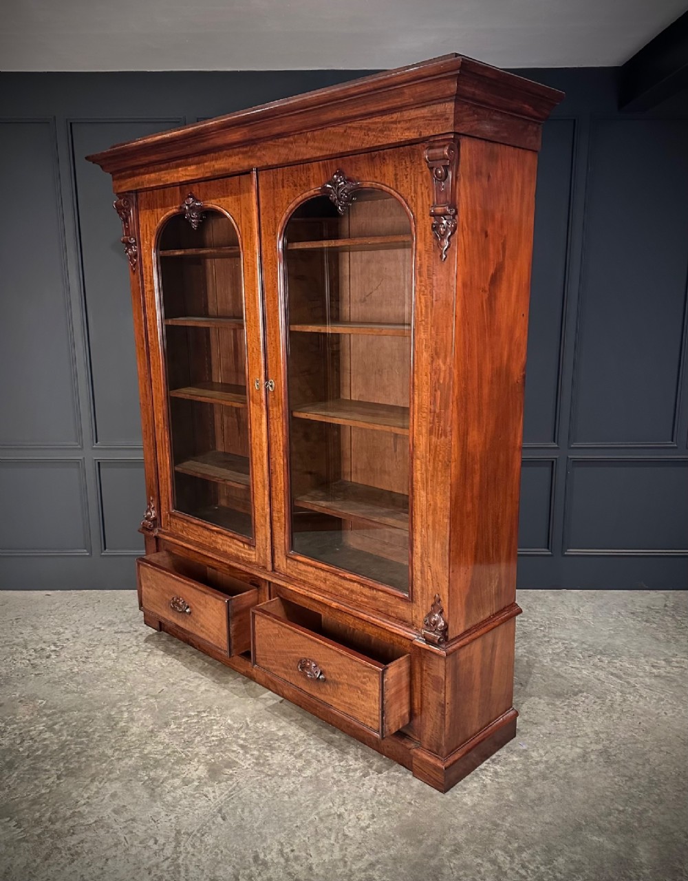 A large Victorian mahogany bookcase featuring two arched glazed doors, carved corbels, internal wooden shelves, and two drawers at the base. View X