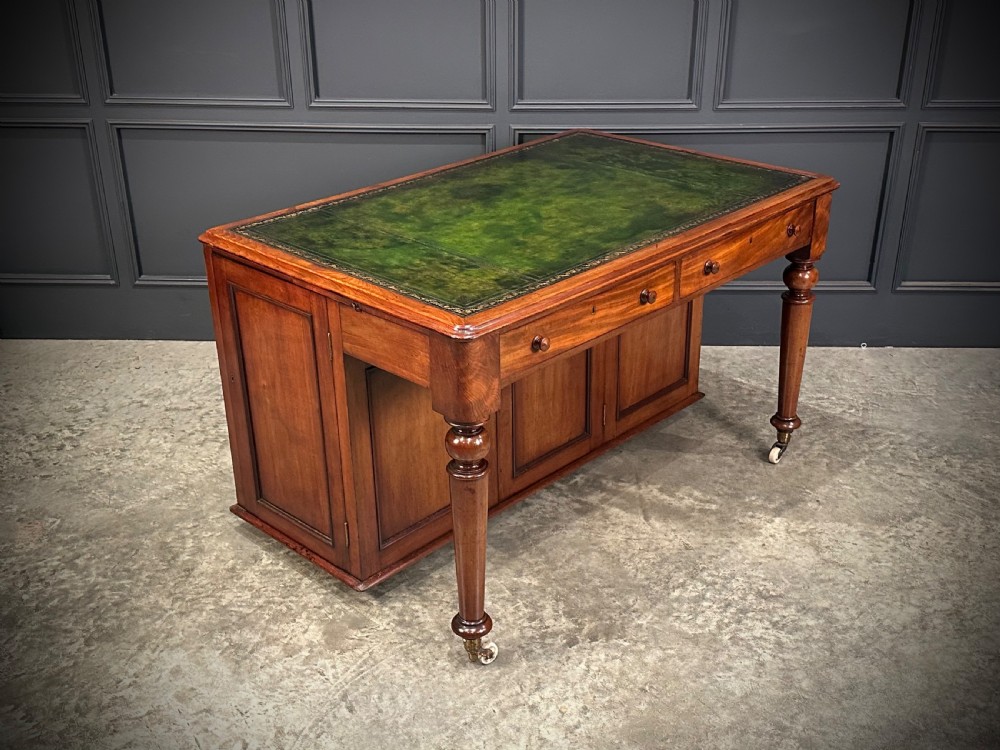 A Victorian mahogany writing desk featuring a green leather inset top, frieze drawers, turned legs on brass castors, and lower panelled cabinet doors.