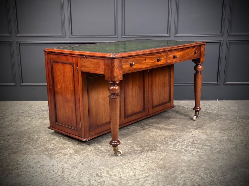 A Victorian mahogany writing desk featuring a green leather inset top, frieze drawers, turned legs on brass castors, and lower panelled cabinet doors. View II