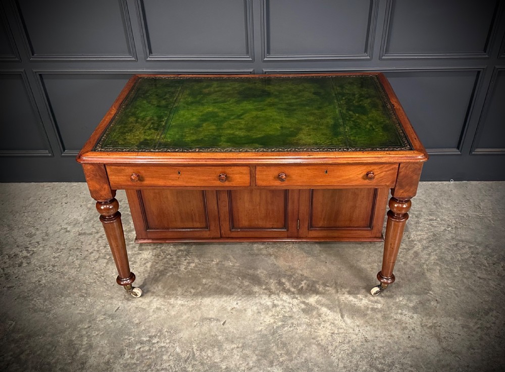 A Victorian mahogany writing desk featuring a green leather inset top, frieze drawers, turned legs on brass castors, and lower panelled cabinet doors. View IV