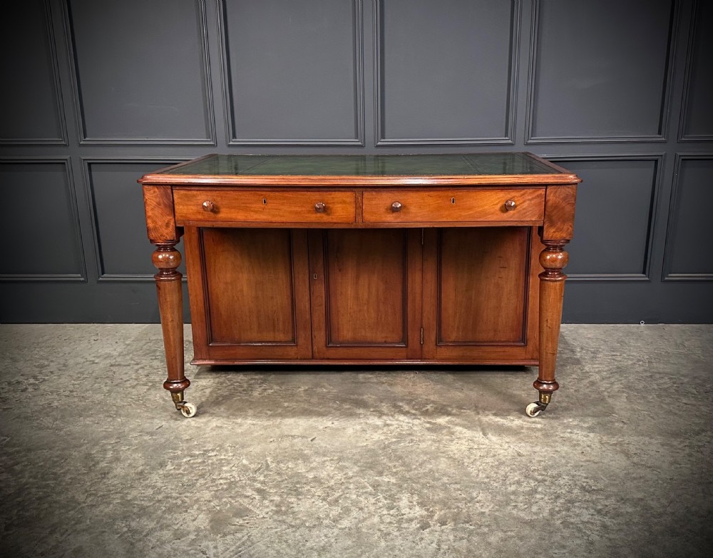 A Victorian mahogany writing desk featuring a green leather inset top, frieze drawers, turned legs on brass castors, and lower panelled cabinet doors. View V