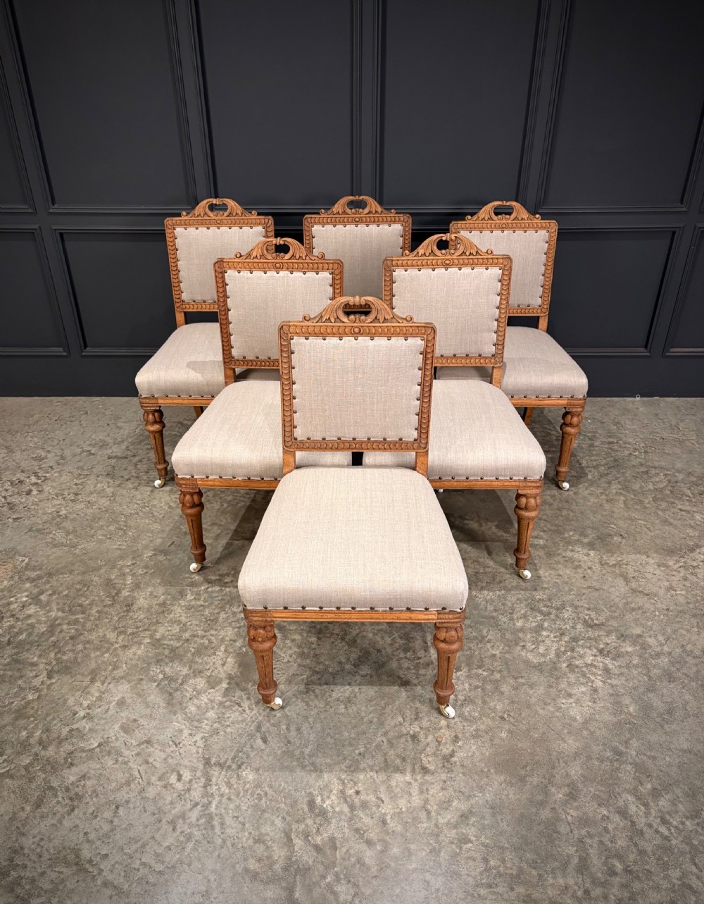 A set of six carved bleached oak dining chairs upholstered in neutral linen fabric with brass stud trimming and turned front legs on ceramic castors. View II