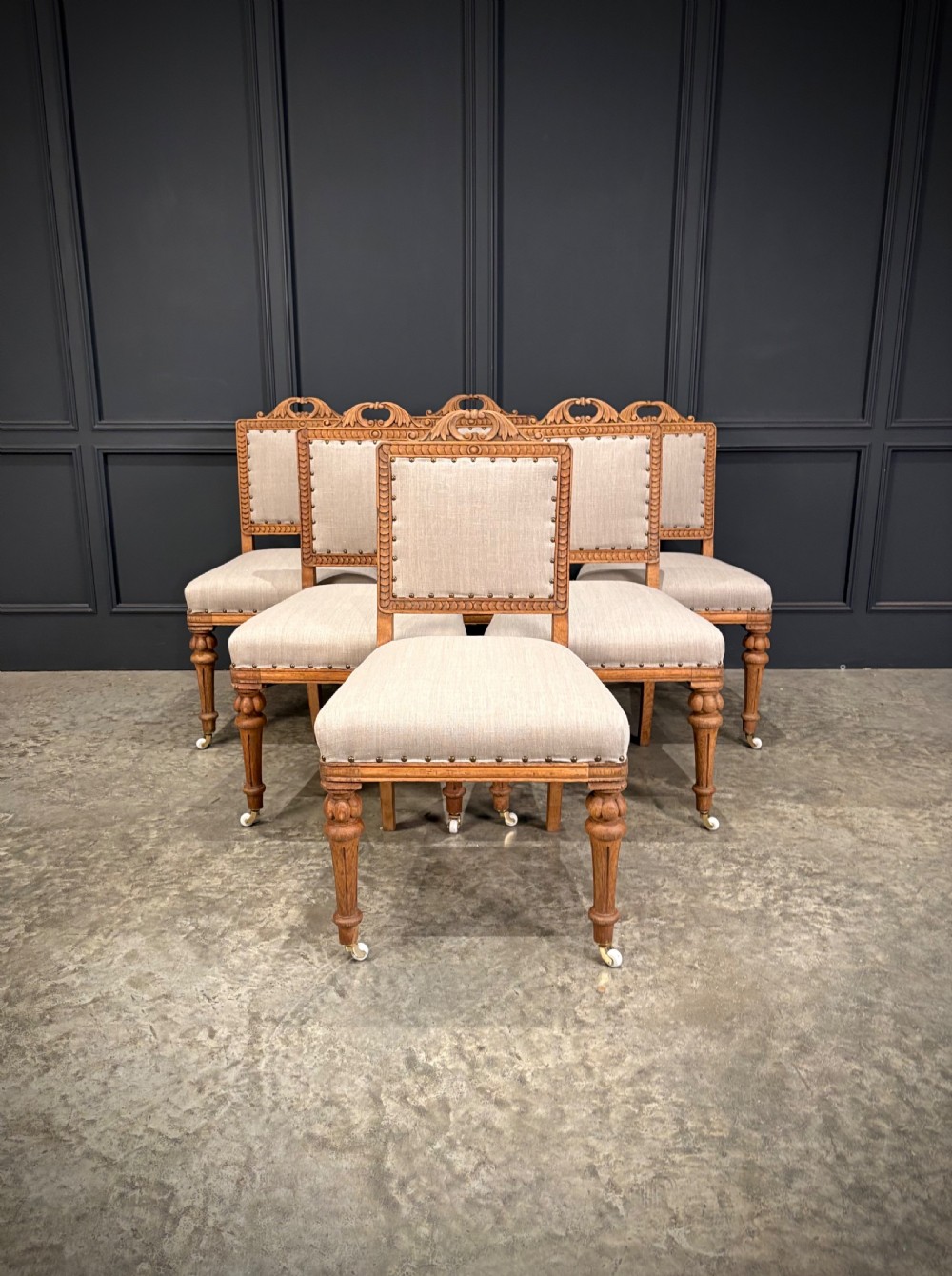 A set of six carved bleached oak dining chairs upholstered in neutral linen fabric with brass stud trimming and turned front legs on ceramic castors.