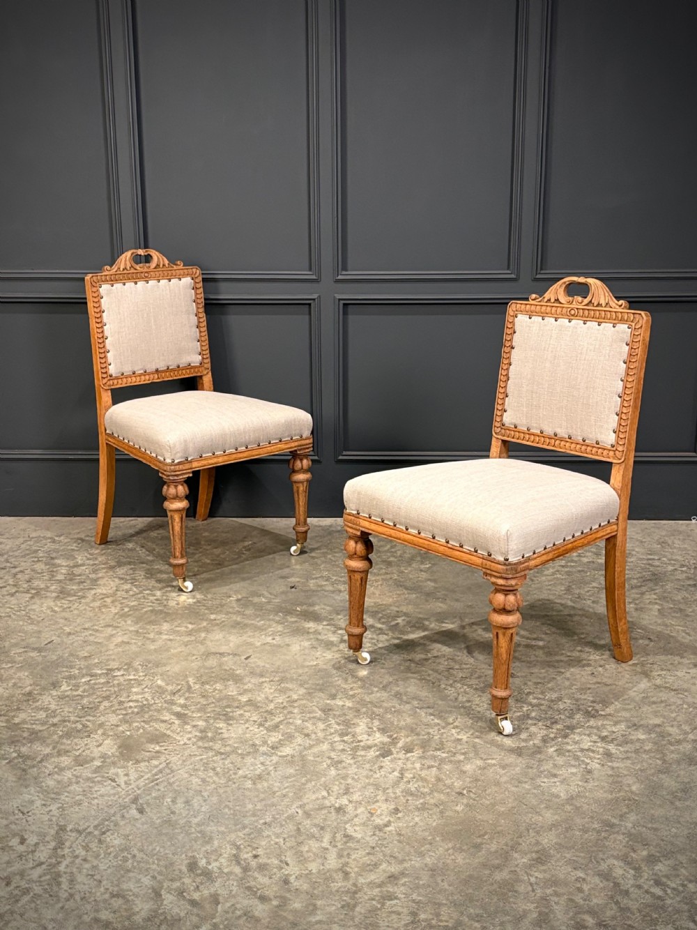 A set of six carved bleached oak dining chairs upholstered in neutral linen fabric with brass stud trimming and turned front legs on ceramic castors. View III