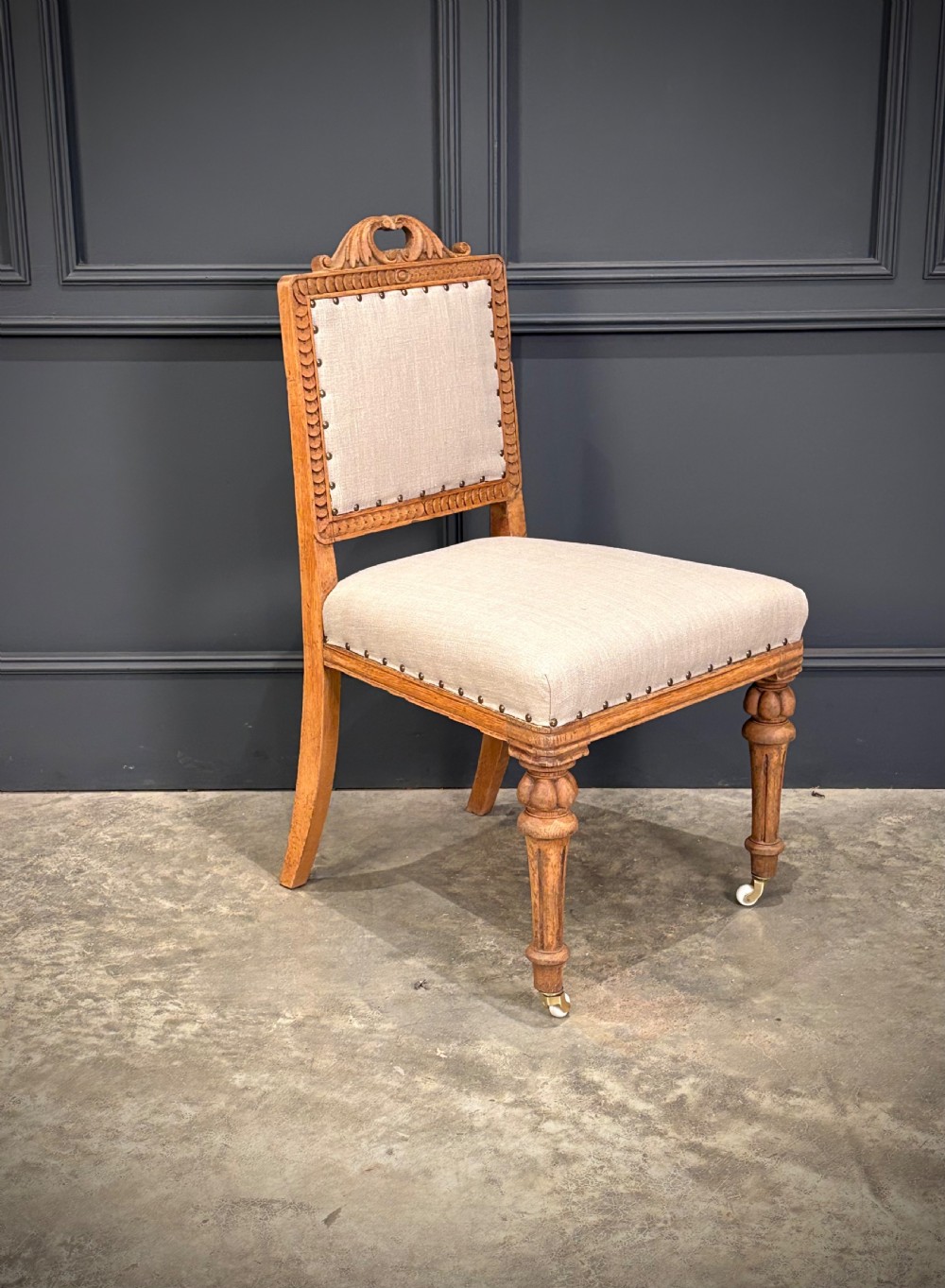 A set of six carved bleached oak dining chairs upholstered in neutral linen fabric with brass stud trimming and turned front legs on ceramic castors. View IV