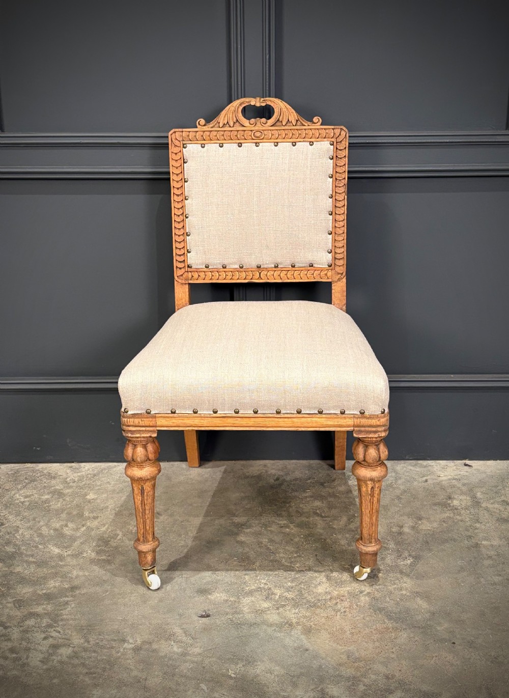 A set of six carved bleached oak dining chairs upholstered in neutral linen fabric with brass stud trimming and turned front legs on ceramic castors. View VI