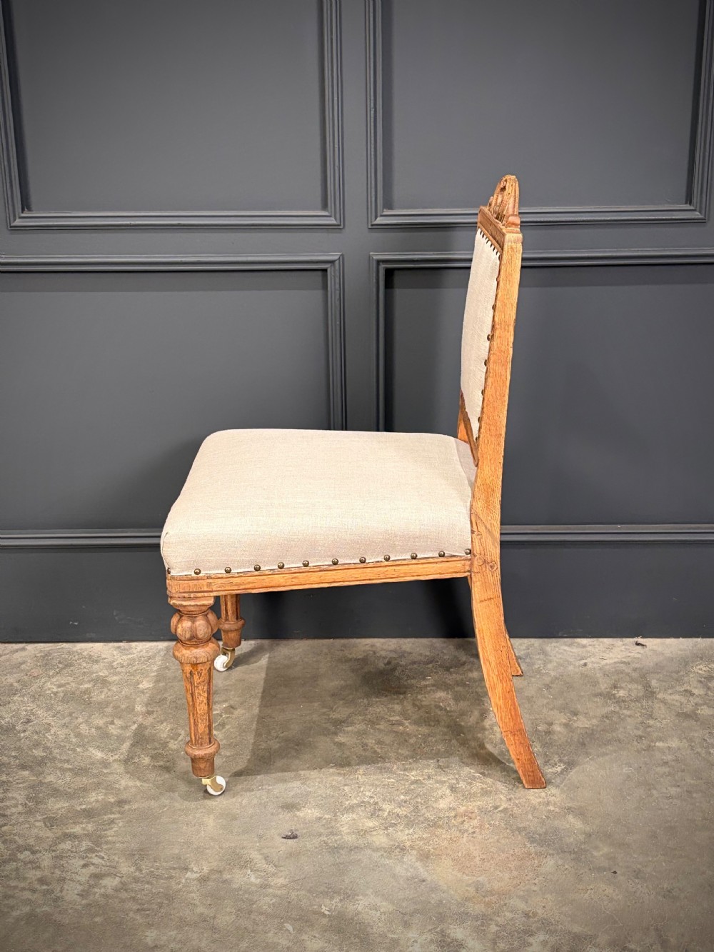A set of six carved bleached oak dining chairs upholstered in neutral linen fabric with brass stud trimming and turned front legs on ceramic castors. View X