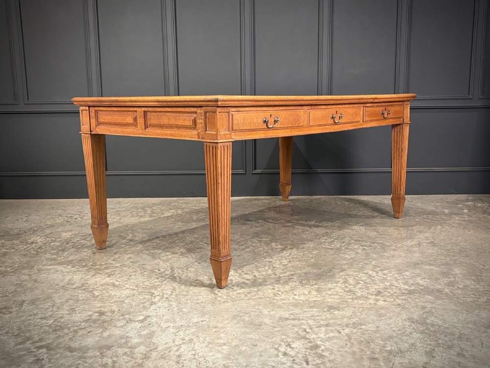 A large oak writing table featuring a green leather top, three frieze drawers with brass drop handles, and four fluted legs. View IV