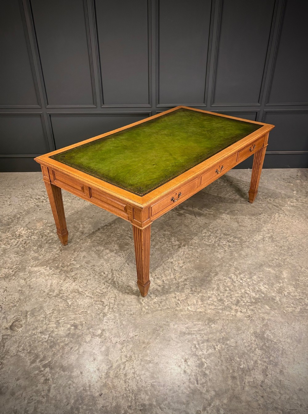 A large oak writing table featuring a green leather top, three frieze drawers with brass drop handles, and four fluted legs. View II