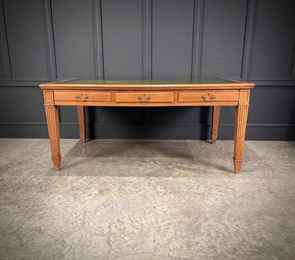 A large oak writing table featuring a green leather top, three frieze drawers with brass drop handles, and four fluted legs. View III