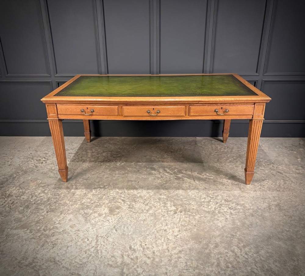 A large oak writing table featuring a green leather top, three frieze drawers with brass drop handles, and four fluted legs.