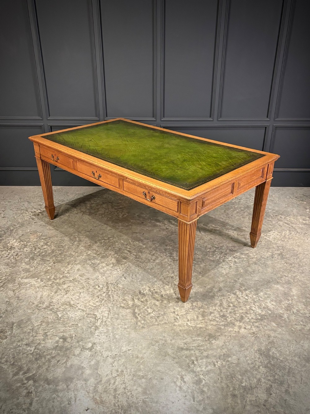 A large oak writing table featuring a green leather top, three frieze drawers with brass drop handles, and four fluted legs. View V