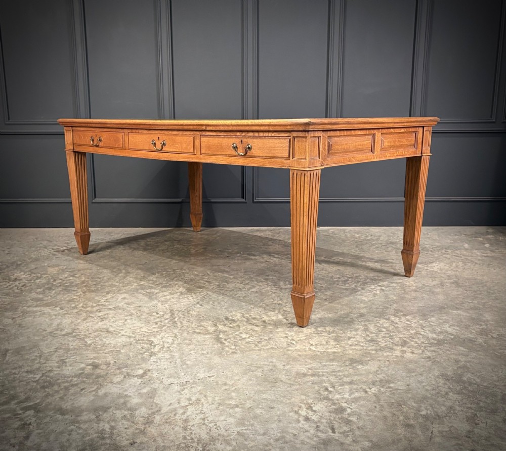 A large oak writing table featuring a green leather top, three frieze drawers with brass drop handles, and four fluted legs. View VI