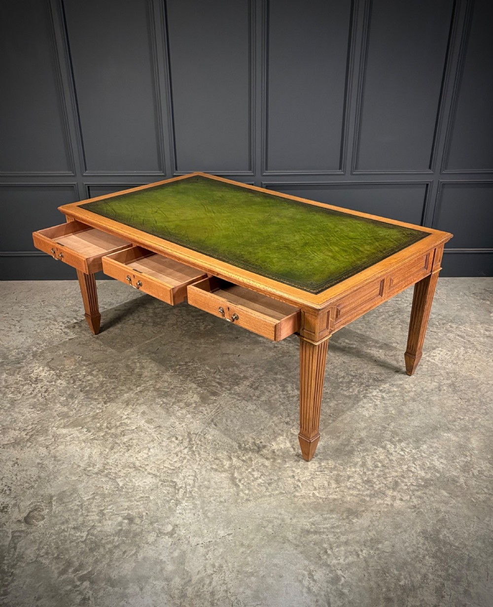 A large oak writing table featuring a green leather top, three frieze drawers with brass drop handles, and four fluted legs. View VII