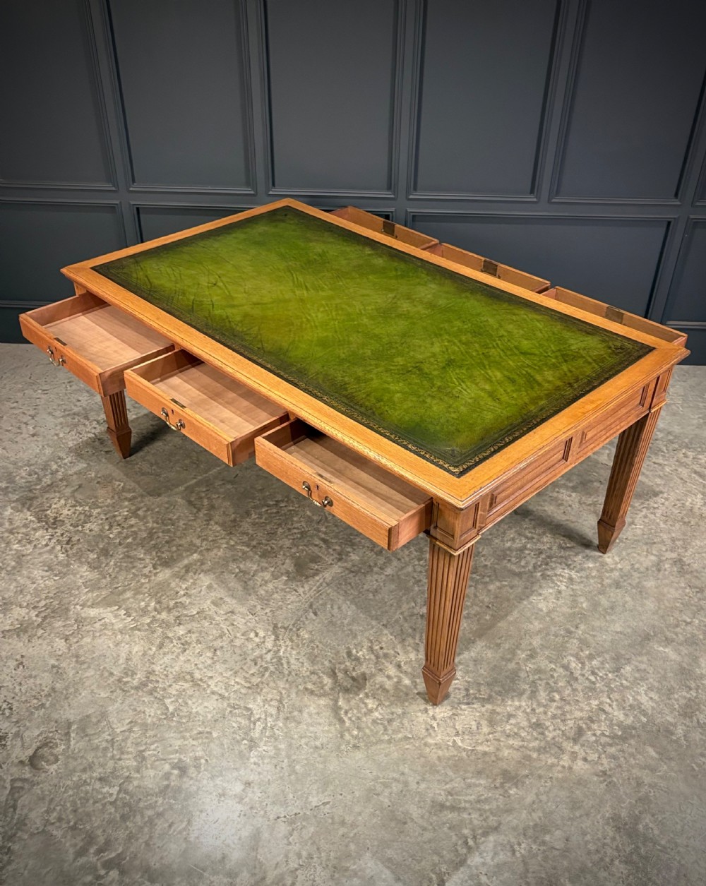 A large oak writing table featuring a green leather top, three frieze drawers with brass drop handles, and four fluted legs. View VIII