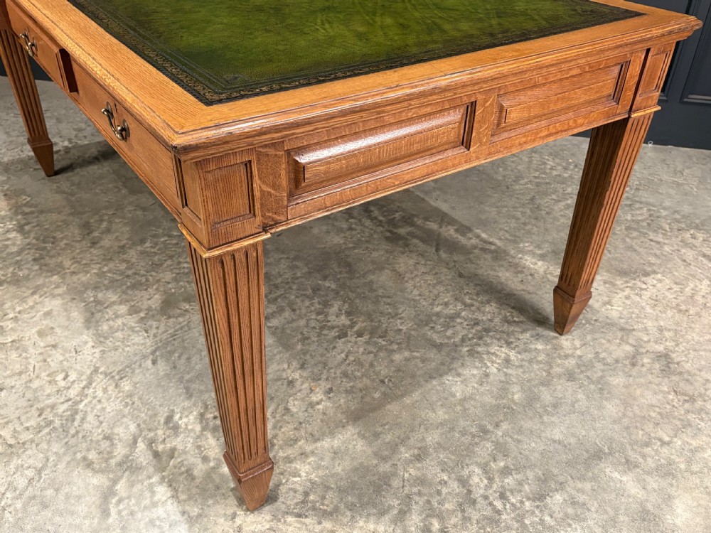 A large oak writing table featuring a green leather top, three frieze drawers with brass drop handles, and four fluted legs. View X