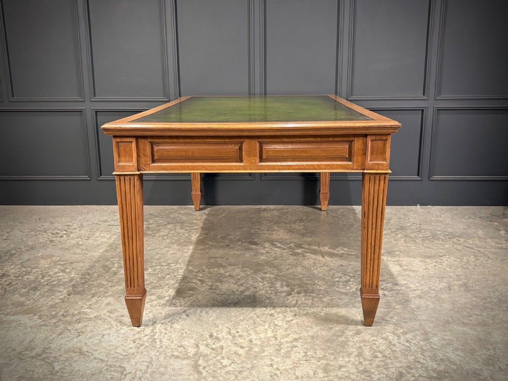 A large oak writing table featuring a green leather top, three frieze drawers with brass drop handles, and four fluted legs. View XI