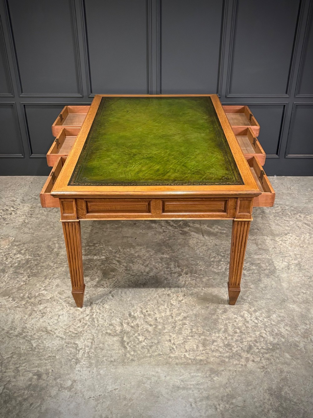 A large oak writing table featuring a green leather top, three frieze drawers with brass drop handles, and four fluted legs. View XII