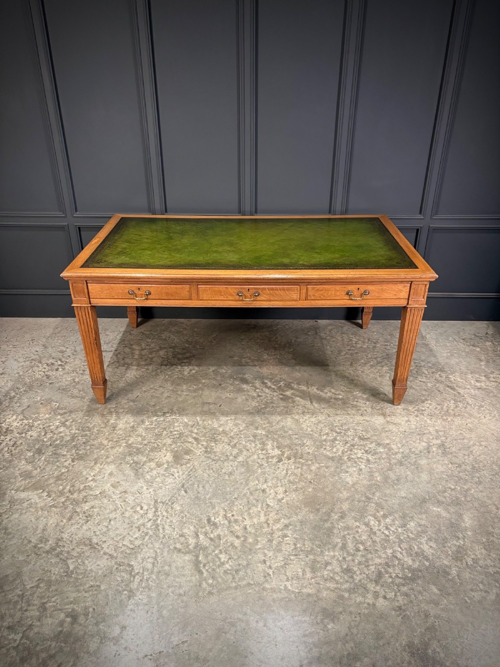 A large oak writing table featuring a green leather top, three frieze drawers with brass drop handles, and four fluted legs. View XIII